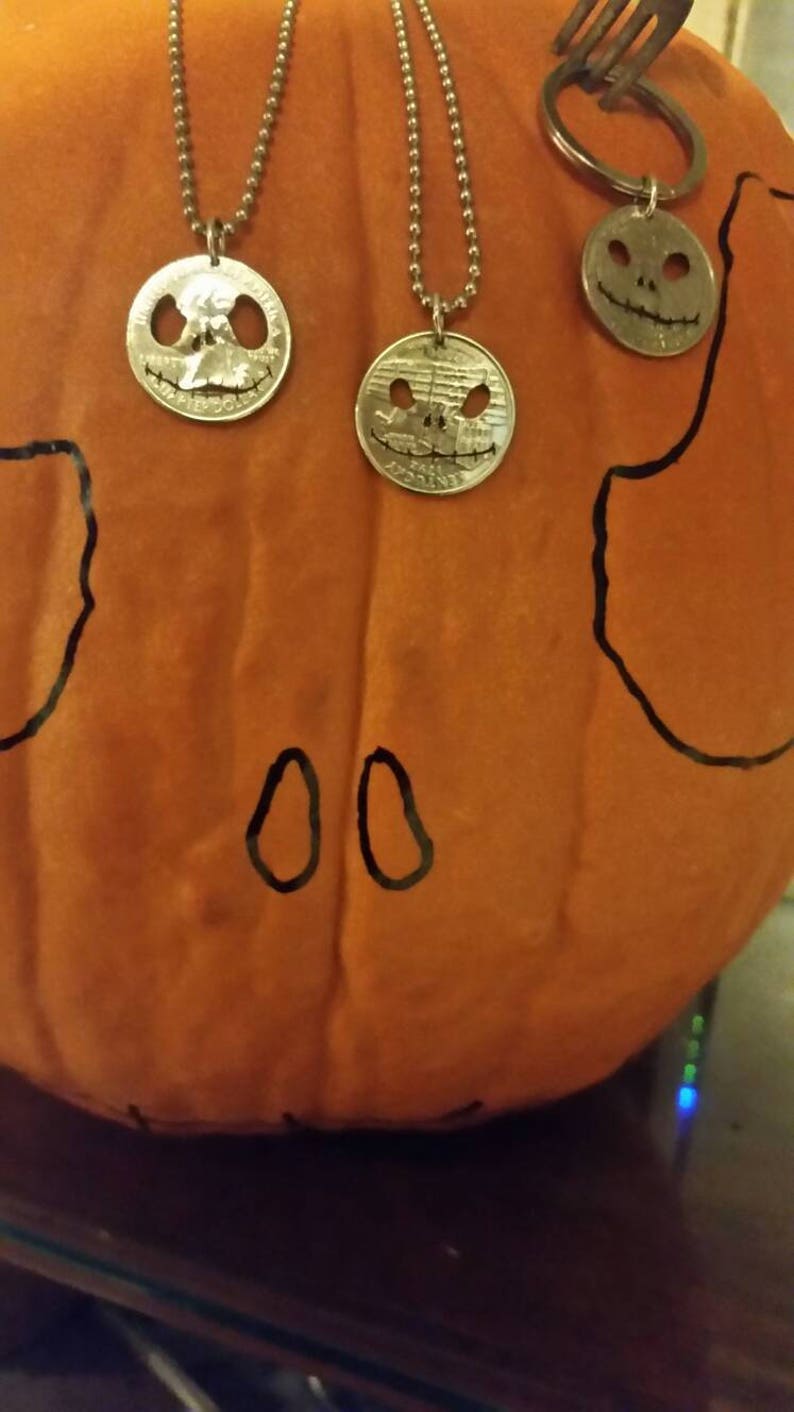 May include: Three silver coin pendants with carved faces, hanging on ball chains, and a keychain with a similar coin. The pendants and keychain are displayed on an orange pumpkin with black drawn features.
