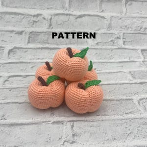 Peut inclure: Cinq pêches crochetées de couleur orange clair, chacune avec une tige marron et une feuille verte. Le mot "PATTERN" est imprimé en noir au-dessus des pêches sur un fond de mur de briques blanches.