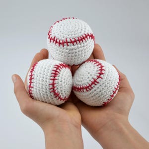 Può includere: Tre palline da baseball bianche all'uncinetto con cuciture rosse sono tenute in mani a coppa su uno sfondo bianco. Le palline da baseball sono rotonde e presentano il classico motivo a punto. Un oggetto artigianale.
