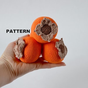 Può includere: Tre cachi arancioni all'uncinetto con cime a forma di fiore marrone sono tenuti in una mano. La parola "PATTERN" è stampata in nero sull'immagine. I cachi sono di un arancione vibrante e le cime dei fiori sono di un marrone a contrasto.