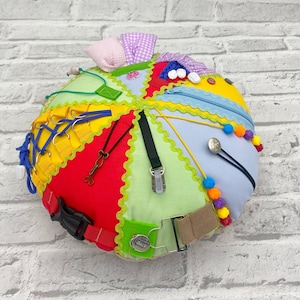 Puede incluir: Una colorida pelota sensorial con varias texturas y cierres. La pelota presenta paneles de tela roja, amarilla, verde y azul, con cremalleras, hebillas y cordones. Incluye una variedad de elementos táctiles, como botones, cintas y pequeños trozos de tela.