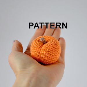 Puede incluir: Un melocotón naranja tejido a mano con un pequeño tallo marrón, sostenido en una mano. La palabra "PATTERN" se muestra en texto negro encima del melocotón. Un objeto artesanal, probablemente decorativo.