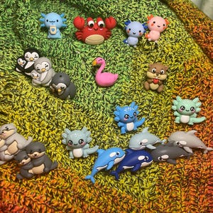 Könnte beinhalten: Eine Sammlung farbenfroher, miniaturisierter Tierfiguren, darunter Pinguine, Otter, Axolotl, Krabben, Delfine und ein Flamingo, angeordnet auf einer strukturierten, mehrfarbigen Oberfläche. Die Figuren bestehen hauptsächlich aus Kunstharz.