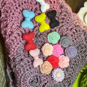 Könnte beinhalten: Eine Sammlung farbenfroher Silikonverzierungen, darunter Schleifen in Weiß, Blau, Gelb, Schwarz, Rot und Rosa, sowie Rosen- und Gänseblümchenformen. Sie sind auf einer rosa-grauen Häkeloberfläche angeordnet.