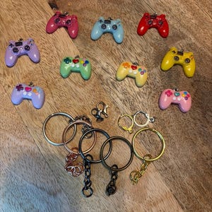 Könnte beinhalten: Eine Sammlung kleiner, bunter Videospiel-Controller-Anhänger in den Farben Pink, Blau, Rot, Lila, Grün, Gelb und Hellrosa, zusammen mit verschiedenen Schlüsselringen, Karabinerverschlüssen und Ohrringhaken in Silber-, Gold- und Bronzetönen.