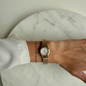 May include: A gold-toned wristwatch with a mesh band and a small, round face. The watch is paired with a delicate gold bracelet. The watch face displays the time with black numerals and hands. The watch and bracelet are worn on a wrist.