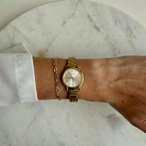 Peut inclure: Une montre dorée avec un cadran rond et un bracelet doré assorti. La montre a un cadran de couleur claire avec des marqueurs et des aiguilles dorés. Le bracelet présente un design de chaîne délicate. La montre et le bracelet sont portés au poignet.