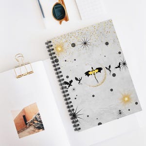 Op de afbeelding: Spiraalgebonden notitieboek met een lichtgrijze kaft met een drakenontwerp. Het ontwerp bevat zwarte drakensilhouetten, gouden glitter en sterren. Het notitieboek ligt open op een wit oppervlak.