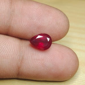 May include: A pear-shaped, faceted ruby gemstone. The gem is a deep red color with bright white facets. The ruby is held between two fingers, showcasing its size and brilliance. This gemstone is ideal for jewelry making.