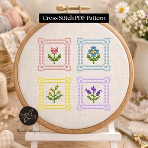 May include: A cross-stitch PDF pattern displayed in a wooden embroidery hoop. The pattern features four floral designs in pink, blue, yellow, and purple, each within a square frame. The text "Cross Stitch PDF Pattern" is at the top.