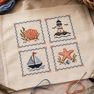 May include: A square piece of off-white Aida fabric featuring four cross-stitch designs arranged in a grid. The motifs include a peach seashell, a lighthouse on a rock, a blue and white sailboat, and a peach starfish, all framed by blue borders.