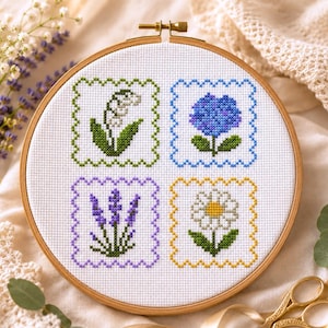 Könnte beinhalten: Ein runder Holzstickrahmen mit weißem Stoff, der vier im Kreuzstich gestickte Blumenquadrate zeigt. Die Motive umfassen ein weißes Maiglöckchen, eine blaue Hortensie, violetten Lavendel und ein weißes Gänseblümchen, jeweils mit farbigem Rand.