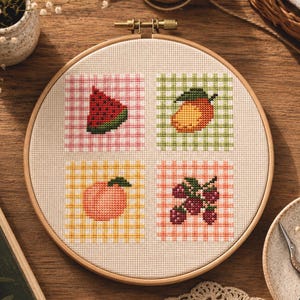 Könnte beinhalten: Ein runder Stickrahmen aus Holz mit einem weißen Aida-Stoff, der vier Kreuzstich-Fruchtmotive zeigt. Die Muster umfassen ein Wassermelonenstück, eine Mango, einen Pfirsich und eine Himbeertraube, jeweils vor einem karierten Hintergrund.