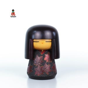 Può includere: Una bambola Kokeshi in legno con una testa arrotondata marrone scuro e capelli neri. Il viso della bambola è dipinto con occhi chiusi e una piccola bocca rossa. Il corpo è decorato con un motivo di foglie d'acero rosse su sfondo nero. La parola "Tamashi" è stampata in alto a sinistra.