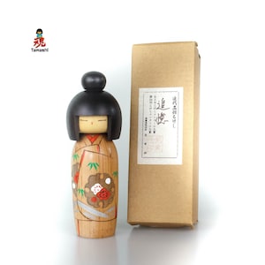 Könnte beinhalten: Eine japanische Kokeshi-Puppe aus Holz mit schwarzem Kopf und einem Körper im Kimono-Stil. Die Puppe hat ein bemaltes Gesicht und steht neben einer braunen Pappschachtel mit japanischem Text. Der Körper der Puppe weist ein Blumendesign mit roten und weißen Akzenten auf.