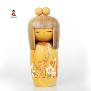 Könnte beinhalten: Eine Kokeshi-Puppe aus Holz mit hellbraunem Bob-Haarschnitt und zwei kleinen Holzkugeln oben. Die Puppe hat ein bemaltes Gesicht mit geschlossenen Augen und ein Blumenmuster in Orange und Weiß. Die Puppe ist von Tamashi.