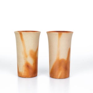 Op de afbeelding: Twee hoge, cilindrische keramische drinkglazen. De glazen hebben een lichtbeige basiskleur met strepen van oranje-bruin. De randen en de onderste delen van de glazen zijn een donkerdere tint oranje-bruin.