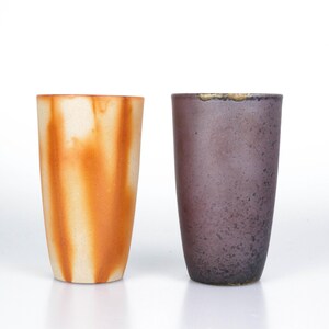 Op de afbeelding: Twee hoge, cilindrische keramische vazen. Eén vaas is oranje en wit met een marmereffect. De andere is donkerpaars met een gespikkelde textuur. Beide vazen hebben een gladde, matte afwerking, tegen een witte achtergrond.