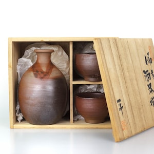 Op de afbeelding: Een Japanse sake-set in een houten kist. De set bevat een bruine keramische fles en twee bijpassende kopjes. De doos heeft Japanse tekst op het deksel. De sake-fles heeft een afgeronde vorm en een smalle hals. De kopjes zijn klein en rond.
