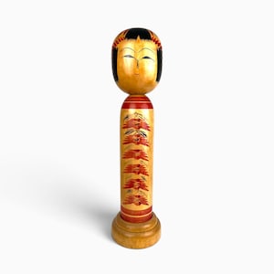 Peut inclure: Une poupée Kokeshi en bois dotée d'une tête arrondie avec des cheveux peints en noir et un visage aux yeux fins et à la petite bouche. Le corps cylindrique est orné d'un motif floral rouge et noir répétitif et repose sur un socle.