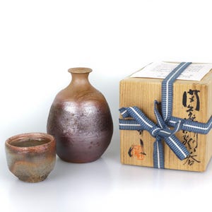 Op de afbeelding: Een keramische sake-set met een bruine, bolvormige fles en een kleine beker. Een houten doos, vastgebonden met een blauw-wit lint, is ook aanwezig. De doos heeft Japanse karakters en een etiket.