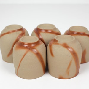 Op de afbeelding: Een set van vijf kleine, cilindrische keramische bekers met een matte beige afwerking en een glad, roodbruin diagonaal glazuurpatroon dat rond de buitenkant van elk vat loopt. De bekers zijn in een groep gerangschikt met de bodems naar boven.
