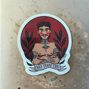 Könnte beinhalten: Ein Aufkleber mit einer stilisierten Illustration einer tätowierten Person mit Schnurrbart und rotem Hintergrund. Der Text "ENBY TRANS EXIST" ist auf einem Banner zu sehen. Der Name des Künstlers, "tylerklee", ist ebenfalls sichtbar.