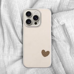 May include: A beige phone case with a brown heart-shaped design. The case has a camera module with three lenses and a flash. The phone case is lying on a white, textured fabric.