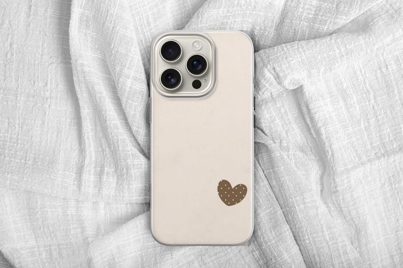 May include: A beige phone case with a brown heart-shaped design. The case has a camera module with three lenses and a flash. The phone case is lying on a white, textured fabric.