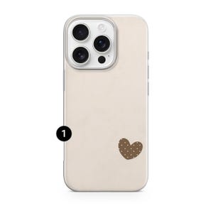 May include: A beige phone case with a brown heart-shaped design featuring white polka dots. The case has a clear border and a camera module with three lenses and a flash. The phone case is designed to protect the phone.