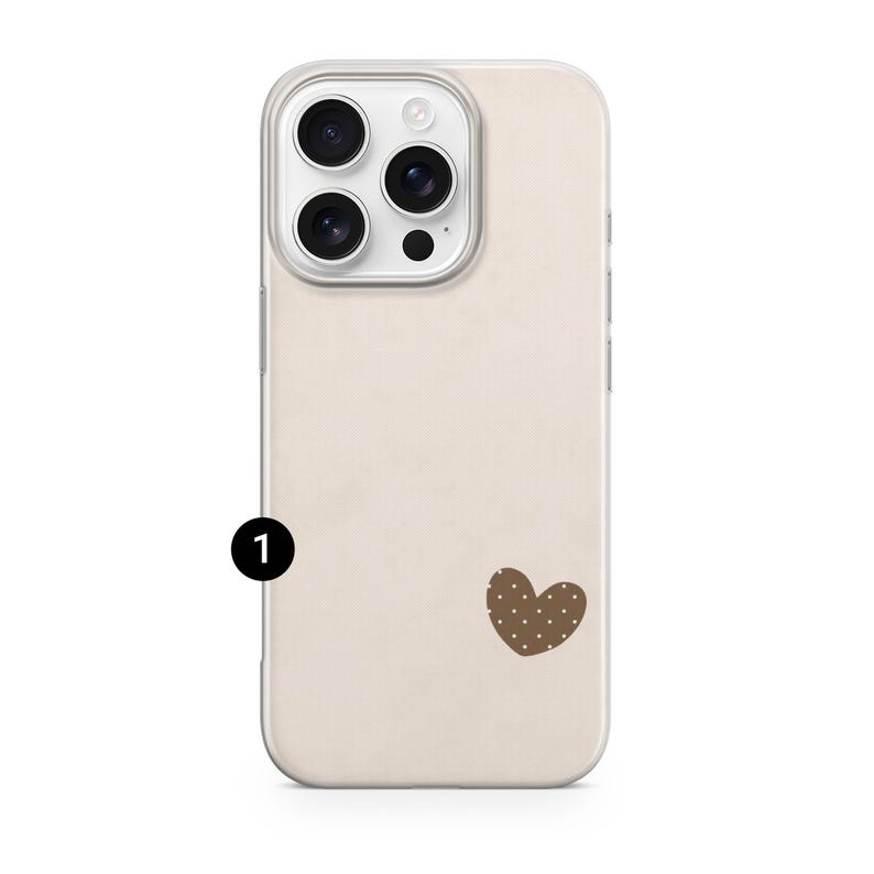 May include: A beige phone case with a brown heart-shaped design featuring white polka dots. The case has a clear border and a camera module with three lenses and a flash. The phone case is designed to protect the phone.