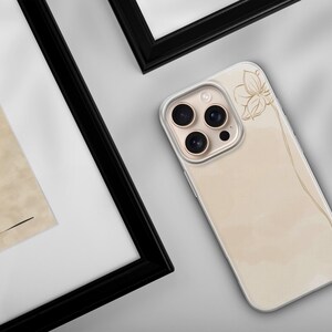 May include: A beige phone case with a floral design and a gold camera lens. The case is lying on a white surface next to a framed artwork with a black frame. The artwork features a profile and a vase.