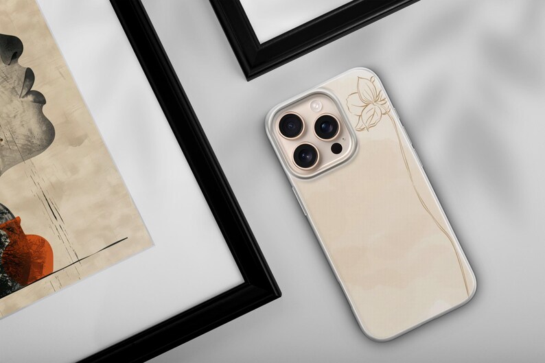 May include: A beige phone case with a floral design and a gold camera lens. The case is lying on a white surface next to a framed artwork with a black frame. The artwork features a profile and a vase.