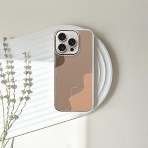 May include: A phone case with a minimalist design featuring abstract shapes in shades of brown and beige. The case is on a white plate, with dried flowers to the left. The phone case has a clear border.