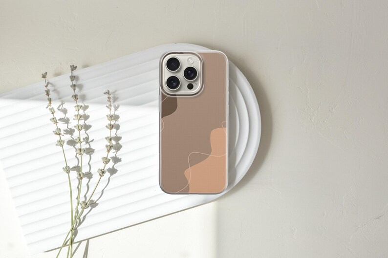May include: A phone case with a minimalist design featuring abstract shapes in shades of brown and beige. The case is on a white plate, with dried flowers to the left. The phone case has a clear border.