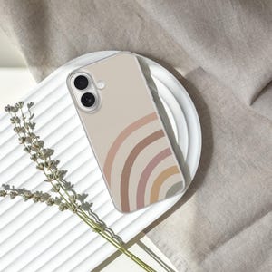 May include: A clear phone case featuring a rainbow design in shades of beige, pink, and brown. The case is resting on a white plate, with a sprig of dried flowers and a neutral-colored fabric in the background.