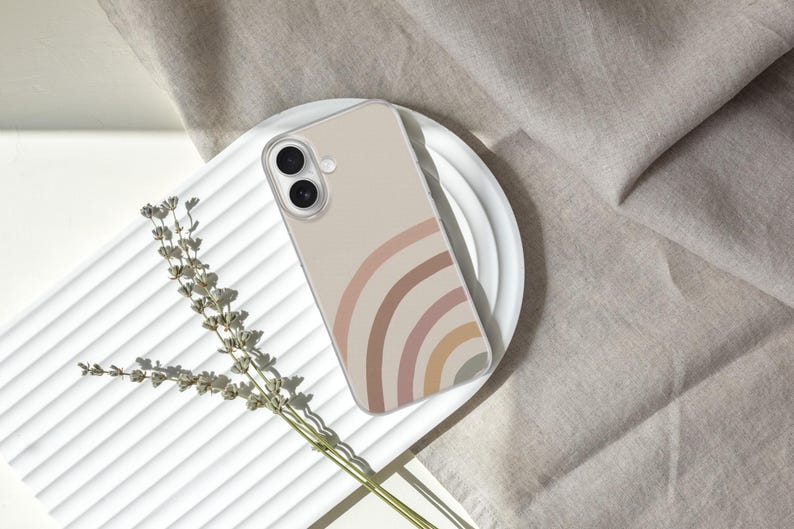 May include: A clear phone case featuring a rainbow design in shades of beige, pink, and brown. The case is resting on a white plate, with a sprig of dried flowers and a neutral-colored fabric in the background.