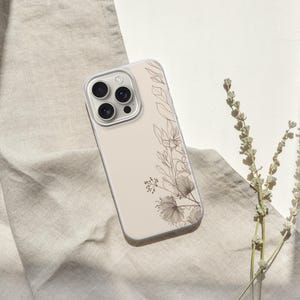 May include: A beige phone case with a floral design featuring delicate line art of leaves and flowers. The case rests on a beige linen cloth, with a sprig of dried flowers to the right.
