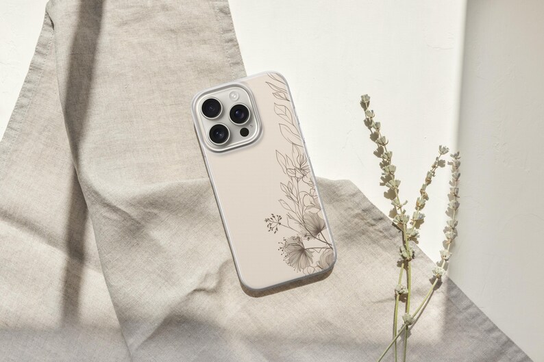 May include: A beige phone case with a floral design featuring delicate line art of leaves and flowers. The case rests on a beige linen cloth, with a sprig of dried flowers to the right.