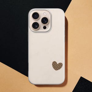 May include: A cream-colored phone case with a brown heart-shaped design featuring white polka dots. The case has a camera module with three lenses and a flash. The background is a combination of black and tan.