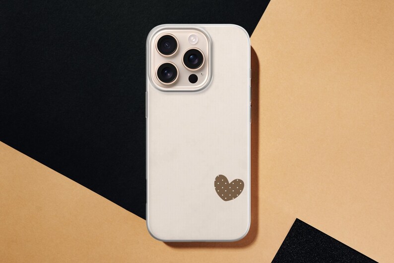 May include: A cream-colored phone case with a brown heart-shaped design featuring white polka dots. The case has a camera module with three lenses and a flash. The background is a combination of black and tan.
