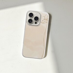 May include: A beige phone case with a floral design. The case has a clear border and a soft, watercolor-like background. A gold-colored flower and a curved line are on the side of the case. The phone case is angled on a white surface.