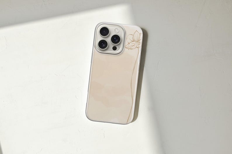 May include: A beige phone case with a floral design. The case has a clear border and a soft, watercolor-like background. A gold-colored flower and a curved line are on the side of the case. The phone case is angled on a white surface.