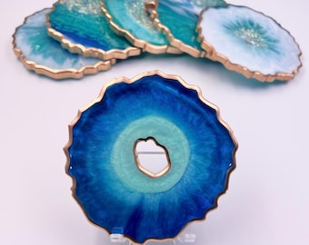 May include: Set of six resin coasters with a geode design. The coasters feature shades of blue and teal with gold edges. One coaster is displayed in the foreground, showcasing the intricate details and color variations. The coasters are designed to protect surfaces from drink rings.