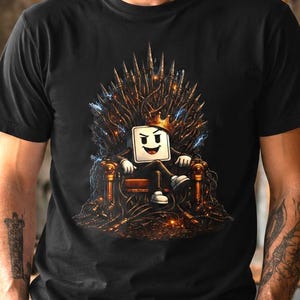 Peut inclure: T-shirt noir avec un personnage de guimauve de dessin animé portant une couronne et assis sur un trône. Le trône présente des détails dorés et bruns. Le t-shirt est porté par une personne dans un atelier avec le texte 'CRÉER COUPER RÉPÉTER'.