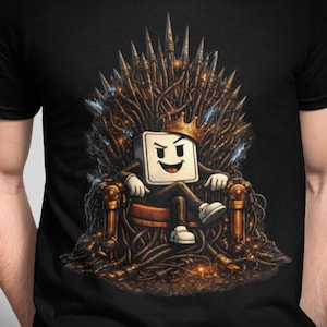 Peut inclure: T-shirt noir avec un personnage de dessin animé couronné, assis sur un trône fait de fils et de pointes. Le personnage est un carré blanc avec un visage souriant, portant un costume et des baskets. Le trône a des accents dorés et bruns.