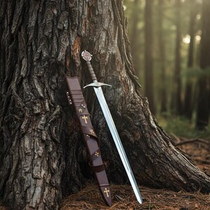 May include: A medieval sword with a silver blade and a brown leather-wrapped handle rests against a tree. A matching brown leather sheath with gold cross accents is next to the sword. The scene is set in a forest.