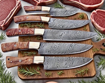 May include: A collection of five Damascus steel knives with wooden handles and brass bolsters, arranged on a wooden cutting board. The blades have a distinctive swirling pattern. Fresh raw steaks and rosemary sprigs surround the knives.