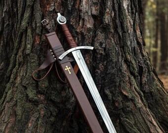 May include: A medieval sword with a silver blade and brown leather-wrapped handle rests against a tree. The sword is in a brown leather sheath with gold cross details. The word "VINCIT" is etched on the blade.