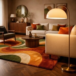 May include: A living room scene with a retro-style rug in shades of orange, green, and cream. A white sofa with orange and green throw pillows sits against a neutral wall. A brass floor lamp with a dome shade illuminates the space, complementing the warm tones.